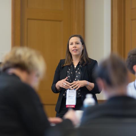 A woman givjing a presentation at the front of a conference room with two large doors.