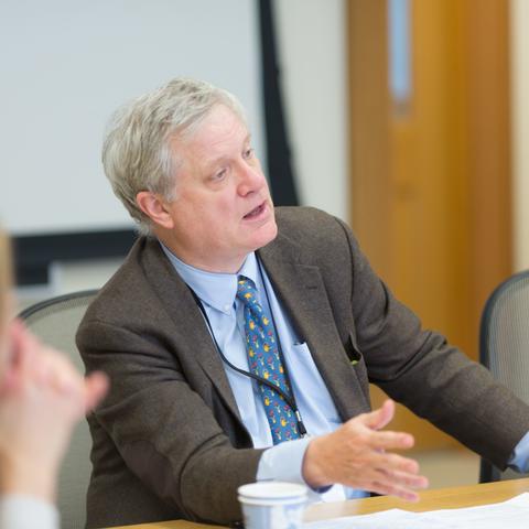 A man with gray hair wearing a brown blazer, a blue tie, and a blue long sleeve button up.