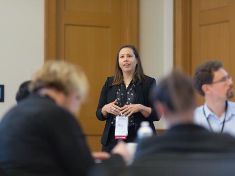 A woman givjing a presentation at the front of a conference room with two large doors.